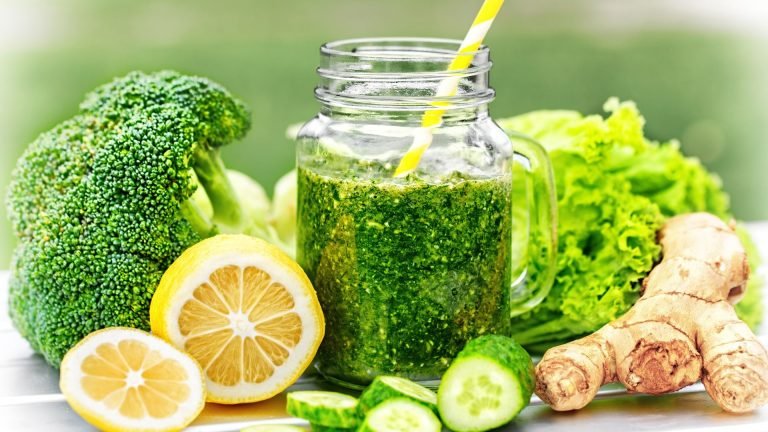 The image showcases a vibrant and healthy green smoothie in a mason jar with a yellow and white striped straw, surrounded by fresh ingredients. Visible are a head of broccoli, half of a lemon with another slice beside it, several cucumber slices, a head of leafy green lettuce, and a piece of ginger root. The colors are vivid, emphasizing the freshness and healthiness of the foods. The background is blurred with shades of green, focusing attention on the jar of smoothie and the raw ingredients. This composition is commonly associated with a healthy lifestyle and nutrition.