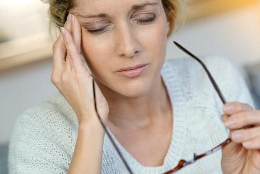 A close-up photograph of a woman who appears to be in discomfort or experiencing a headache. She is holding her glasses in one hand and has her eyes closed, with her other hand pressed to the side of her forehead, indicating pain or stress. She is wearing a light-colored, knitted sweater and has short, wavy blonde hair. The setting seems to be indoors with soft, natural lighting that suggests a calm environment, contrasting with the woman's pained expression.